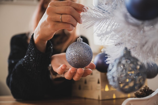 Vista de cerca de las manos femeninas a punto de colgar un adorno navideño azul brillante en el árbol de navidad blanco