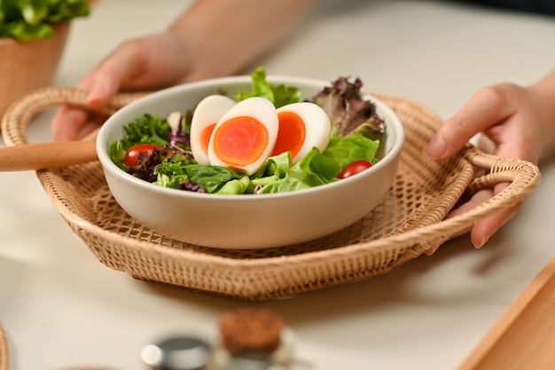 Vista de cerca de manos femeninas sosteniendo una bandeja de mimbre con un plato de ensalada con huevos duros, lechuga y tomate