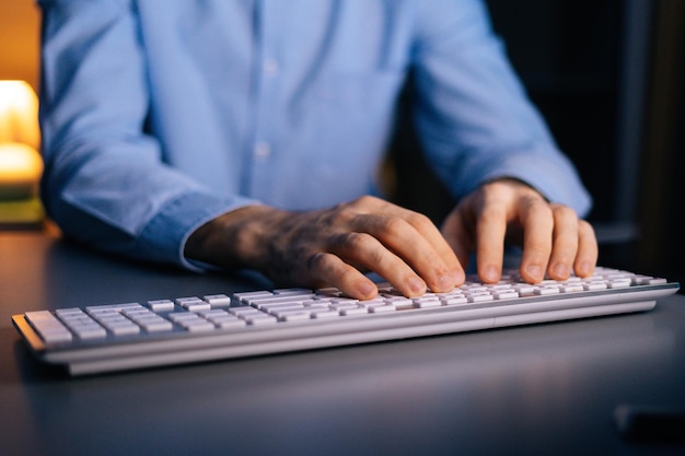 Vista de cerca de las manos de un hombre irreconocible que trabaja escribiendo en una computadora con teclado inalámbrico. El trabajador independiente o estudiante masculino está trabajando sentado en el escritorio de la oficina en casa a altas horas de la noche en una habitación oscura.