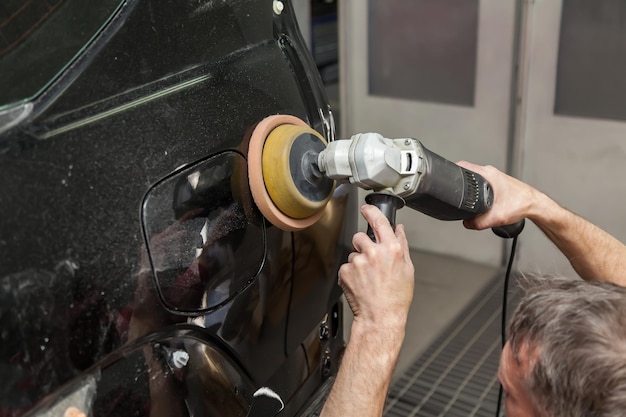 Vista de cerca de las manos de un trabajador masculino que sostiene una herramienta para pulir el guardabarros de un automóvil mientras trabaja en un taller de detalles de vehículos Industria de servicios automotrices