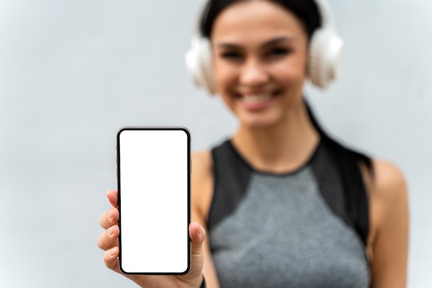 Vista de cerca de la mujer atlética sonriente sosteniendo un teléfono inteligente en el brazo extendido Chica escuchando música sobre fondo de pared gris Enfoque en primer plano