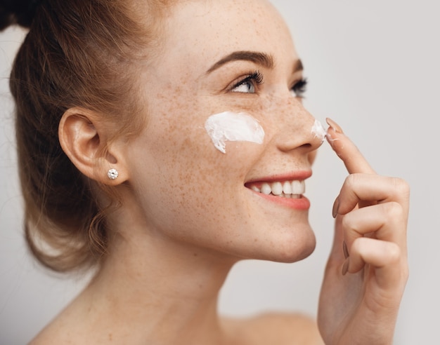 Vista de cerca de una mujer caucásica de pelo rojo con pecas que se aplica una crema en la cara mientras posa a un lado en una pared entera con espacio libre