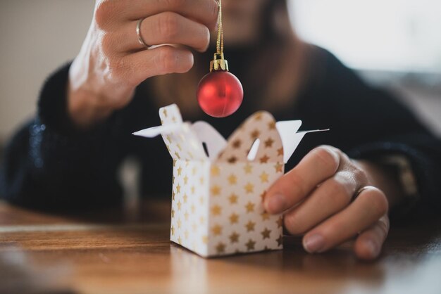 Vista de cerca de una mujer colocando un adorno navideño rojo brillante en una pequeña caja de regalo de Navidad