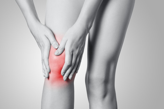 Vista de cerca de una mujer joven con dolor de rodilla sobre fondo gris. Fotografía en blanco y negro con punto rojo.