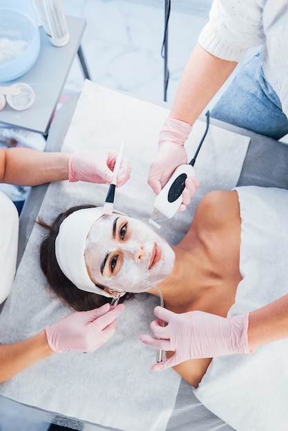 Vista de cerca de la mujer que está acostada en el salón de spa y tiene un procedimiento de limpieza facial mediante diferentes dispositivos y máscara.