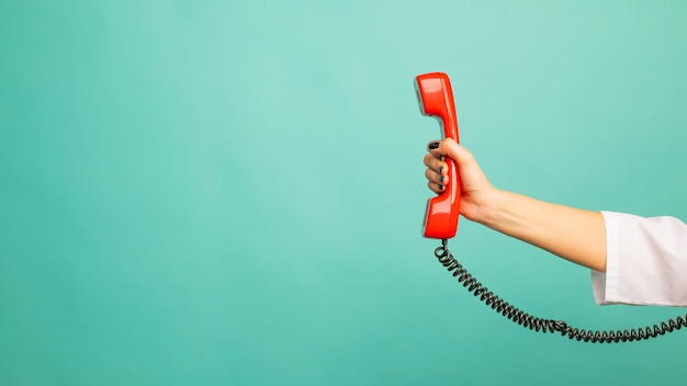 Vista de cerca de la mujer que sostiene el auricular de teléfono con cable rojo sobre fondo azul, espacio para texto. Concepto de línea directa