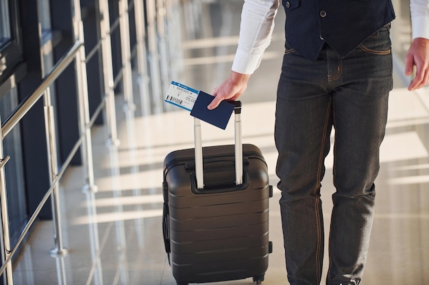 Vista de cerca del pasajero masculino con elegante ropa formal que se encuentra en la sala del aeropuerto con equipaje y boletos.