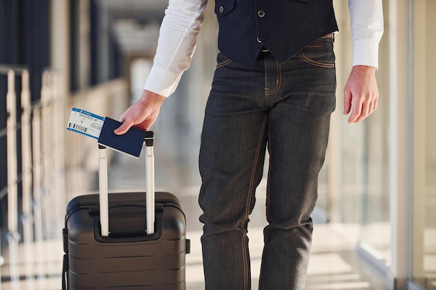 Vista de cerca del pasajero masculino con elegante ropa formal que se encuentra en la sala del aeropuerto con equipaje y boletos.