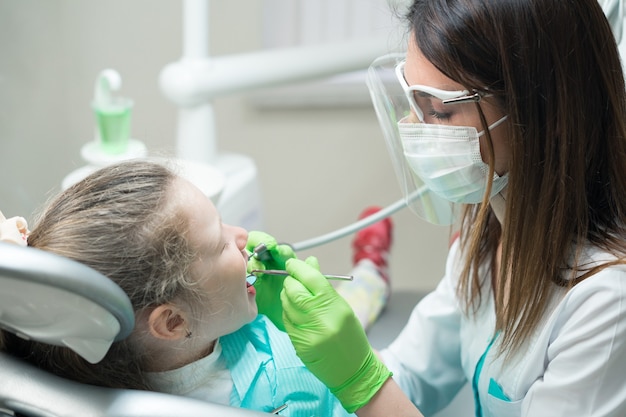 Vista de cerca de la pequeña niña linda en la silla del dentista. dentista en mascarilla tratando los dientes del niño