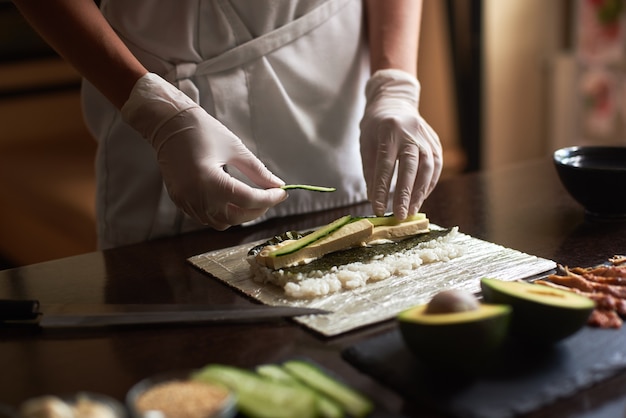 Vista de cerca del proceso de preparación de sushi con nori, arroz, pepino y tortilla en estera de bambú