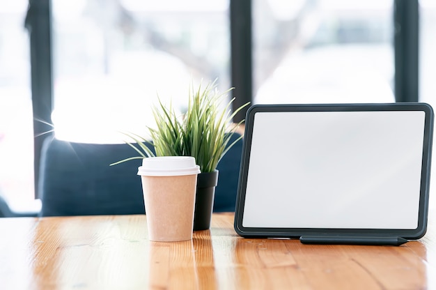 Vista de cerca de la tableta de pantalla en blanco, vaso de papel y planta de interior en la mesa de madera con espacio de copia.