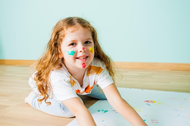 Vista cercana de la cara de una chica encantadora con marcas coloridas. Chica estirándose en el suelo después de pintar con los dedos en su habitación