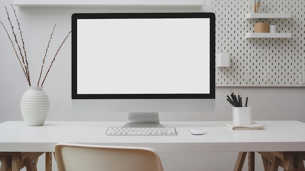 Foto vista cercana del espacio de trabajo con computadora de pantalla en blanco, suministros de oficina, decoración y estante en escritorio blanco con pared blanca