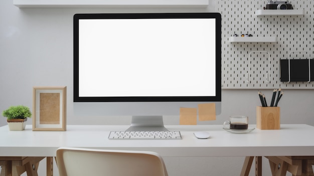 Vista cercana del espacio de trabajo con computadora de pantalla en blanco, suministros de oficina, decoración y estante en escritorio blanco con pared blanca