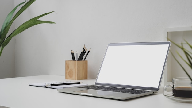 Vista cercana del espacio de trabajo con pantalla en blanco portátil, taza de café y lápices en el escritorio con pared blanca