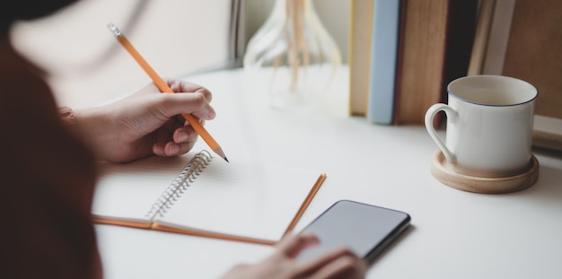 Vista cercana de joven mujer escribiendo su idea en el cuaderno mientras toca su teléfono inteligente