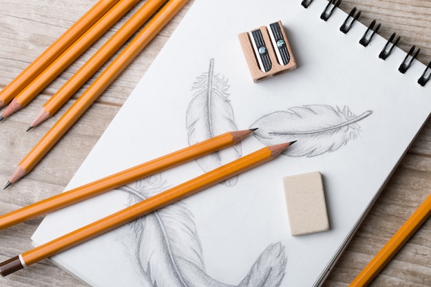 Vista cercana de la mesa del artista o diseñador. Lápices, sacapuntas y borrador en un cuaderno de bocetos con plumas dibujadas a mano