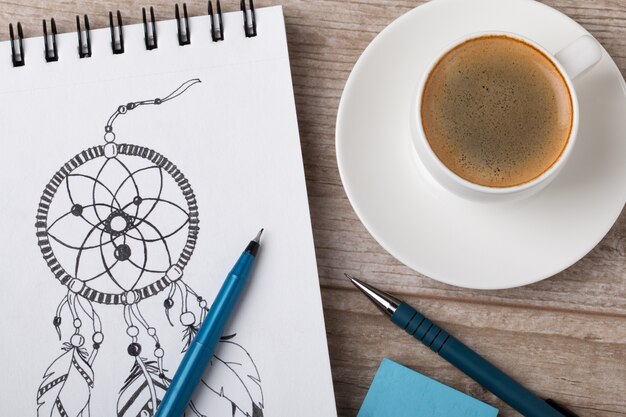 Foto vista cercana de la mesa del artista o diseñador. taza de café, lápiz, delineador fino y borrador en un cuaderno de bocetos con un atrapasueños dibujado a mano