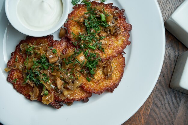 Vista cercana de plato tradicional de la cocina ucraniana - tortitas de patata con crema agria y champiñones en un plato de cerámica blanca