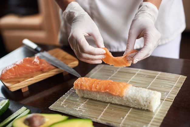 Vista cercana del proceso de preparación de sushi rodante con guantes desechables en estera de bambú