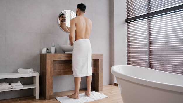 Vista completa árabe atleta musculoso desnudo hombre indio con toalla en las caderas mirando el baño de espejo