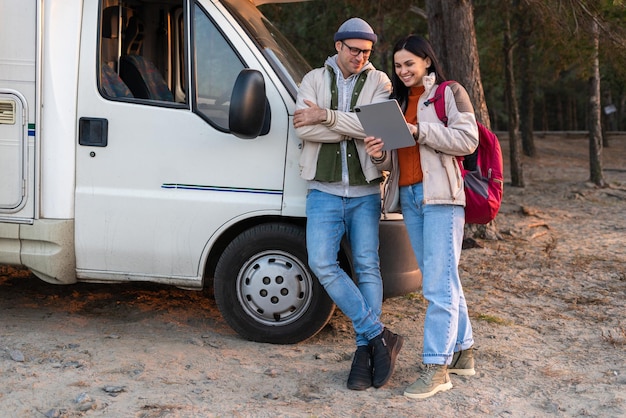 Vista completa de la feliz pareja caucásica usando la aplicación de viaje en la tableta mientras está de pie cerca del auto con el bosque al fondo