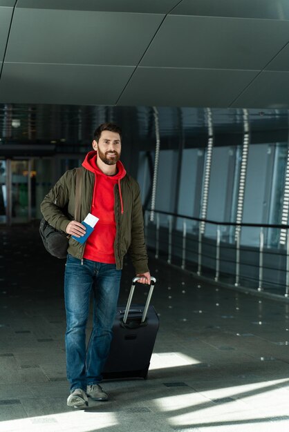 Vista completa del joven seguro de sí mismo que atraviesa el pasillo con una maleta y espera el vuelo en el aeropuerto Concepto de viaje