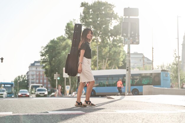 vista completa de una mujer joven con un estuche de guitarra colgado en la espalda caminando por la ciudad