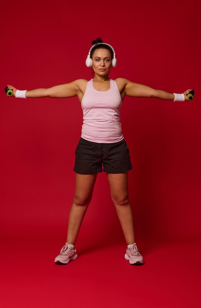 Vista completa del tamaño del cuerpo de una atleta femenina activa haciendo ejercicio con pesas sobre fondo rojo con espacio para copiar texto publicitario