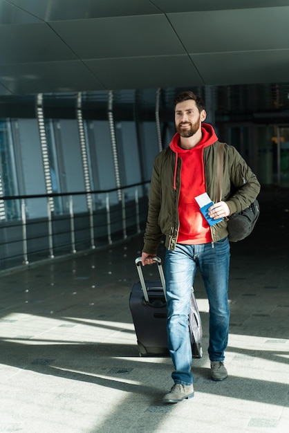 Vista completa del turista masculino tranquilo caminando en el aeropuerto y mirando hacia otro lado con expresión feliz Está sosteniendo su maleta gris