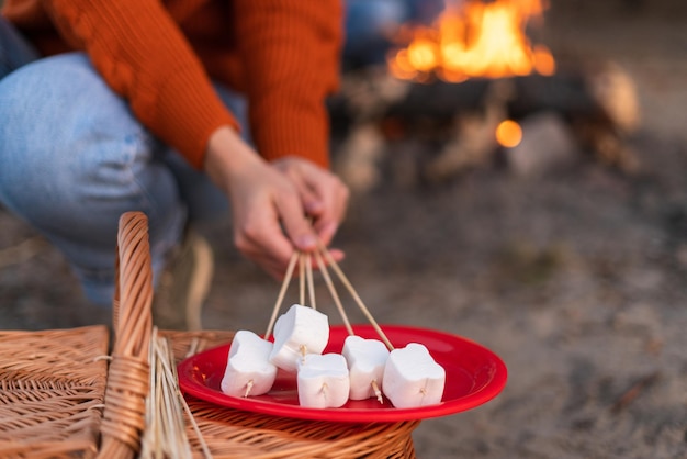 Vista cortada da mulher caucasiana desfrutando de recreação ao ar livre enquanto segura marshmallows nas varas e preparando assado no fogo com o namorado. Foto