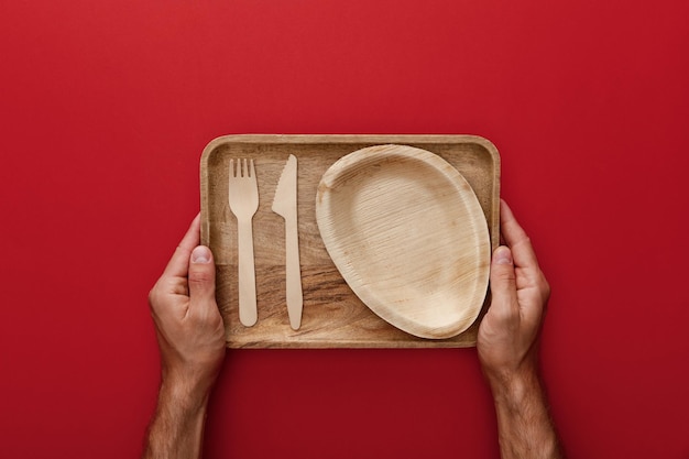 Vista cortada do homem segurando o prato de madeira retangular natural com garfo e faca no vermelho