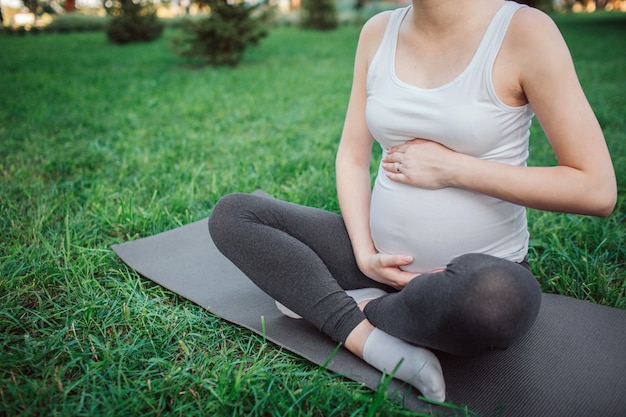 Vista de corte de la joven mujer embarazada sentada en el compañero de yoga en el parque. Ella toma las manos alrededor del vientre. Modelo sentada en postura de loto.