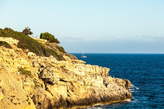 Vista de una costa rocosa y salvaje cerca del mar con personas y un yate a vela en el horizonte