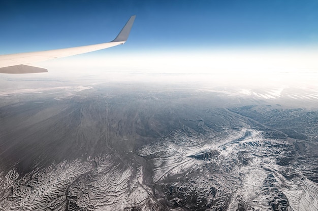 Vista da janela de um avião com asa e terreno dobrado coberto de neve e geleiras nas terras altas iranianas
