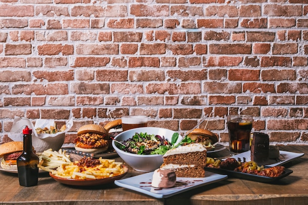 Vista da mesa com uma variedade de pratos, hambúrgueres, batatas fritas e salada, bebidas, asas de frango, molho, bolo e sobremesas na mesa de madeira. Cardápio do restaurante.