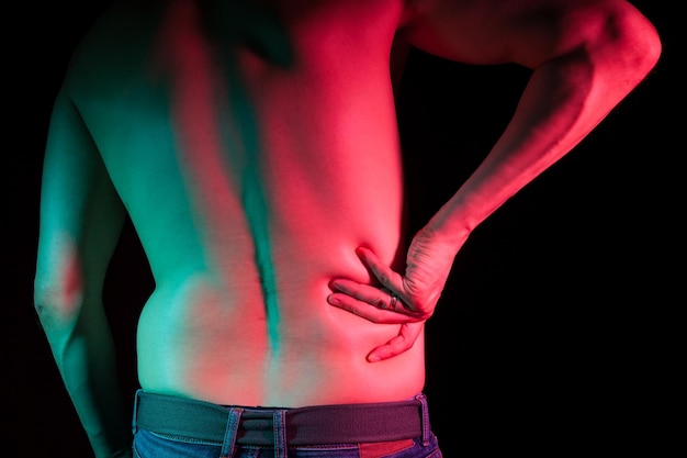 Vista da parte de trás da parte inferior das costas de um homem adulto que segura a mão de lado devido a dor nas costas em um fundo preto com retroiluminação em verde e vermelho medicina e saúde da coluna e do esqueleto