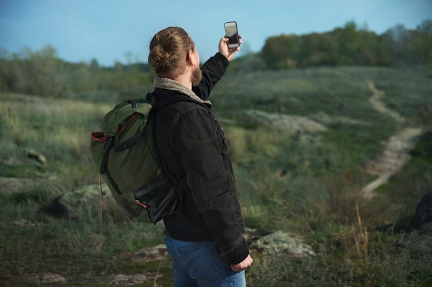 Vista da parte de trás de um caminhante com mochila em busca de sinal em um smartphone, permanecendo em uma passarela nas montanhas