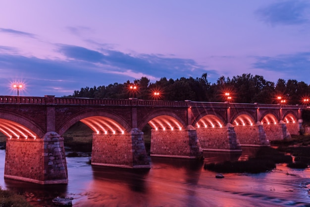 Vista da ponte iluminada sobre o rio contra o céu nublado durante o anoitecer