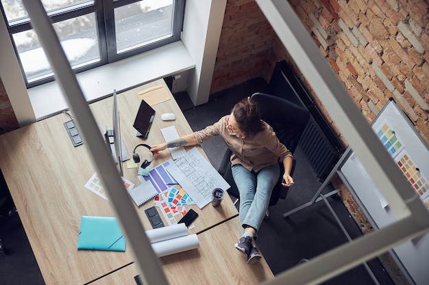 Vista de alto ângulo da designer de interiores feminina pegando fones de ouvido, lendo uma planta enquanto trabalhava