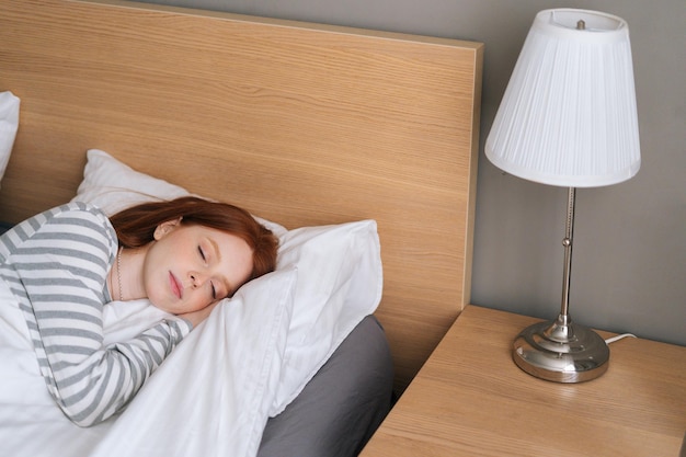 Vista de alto ângulo da jovem ruiva atraente dormindo bem na cama abraçando o cobertor branco macio em casa. Senhora bonita descansando desfrutando de roupa de cama macia fresca e colchão no quarto.