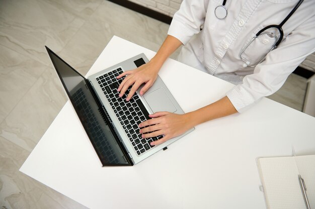 Vista de alto ângulo das mãos da médica, digitando texto no teclado do laptop no consultório médico na clínica branca.