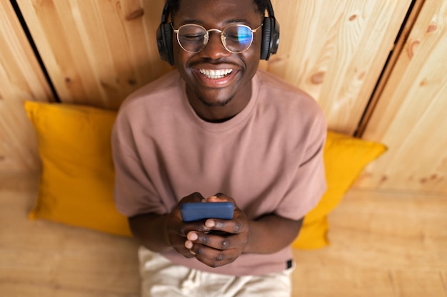 Foto vista de alto ângulo de homem preto ouvindo música com fones de ouvido sem fio usando telefone celular em casa