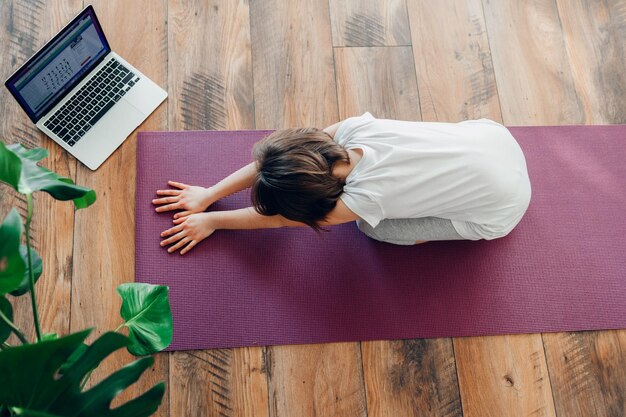 Vista de alto ângulo de menina fazendo ioga em tapete de exercício por laptop