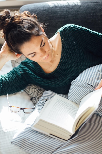 Vista de alto ângulo de mulher jovem lendo um livro enquanto está deitada na cama em casa