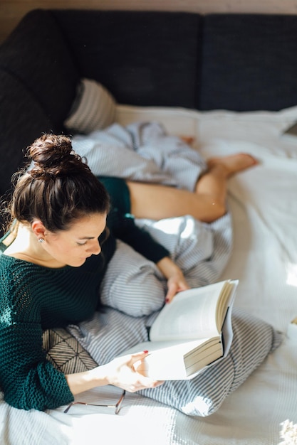 Vista de alto ângulo de mulher jovem lendo um livro enquanto está deitada na cama em casa
