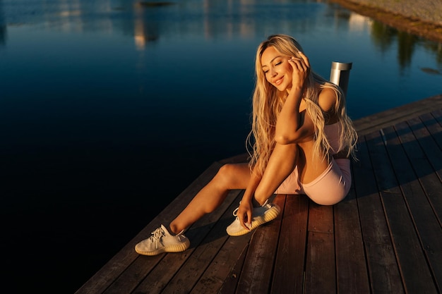 Vista de alto ângulo de mulher loira desportiva alegre em roupas esportivas sentada posando no cais de madeira cite parque pela água na manhã ensolarada de verão olhando para baixo