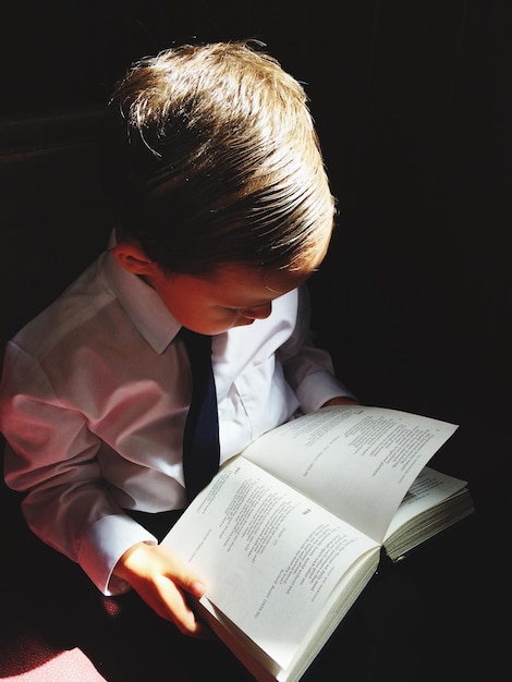 Vista de alto ângulo de um menino lendo um livro à luz do sol na sala escura