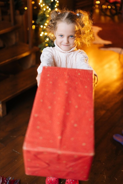 Vista de alto ângulo de uma adorável garotinha loira sorridente enviando caixa de presente com presente de Natal para você olhando para a câmera na sala de estar aconchegante