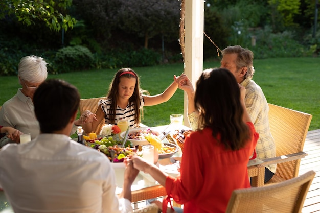 Vista de alto ângulo de uma família caucasiana de várias gerações sentada do lado de fora em uma mesa de jantar preparada para uma refeição, com os olhos fechados, de mãos dadas em oração. Família aproveitando o tempo em casa, conceito de estilo de vida